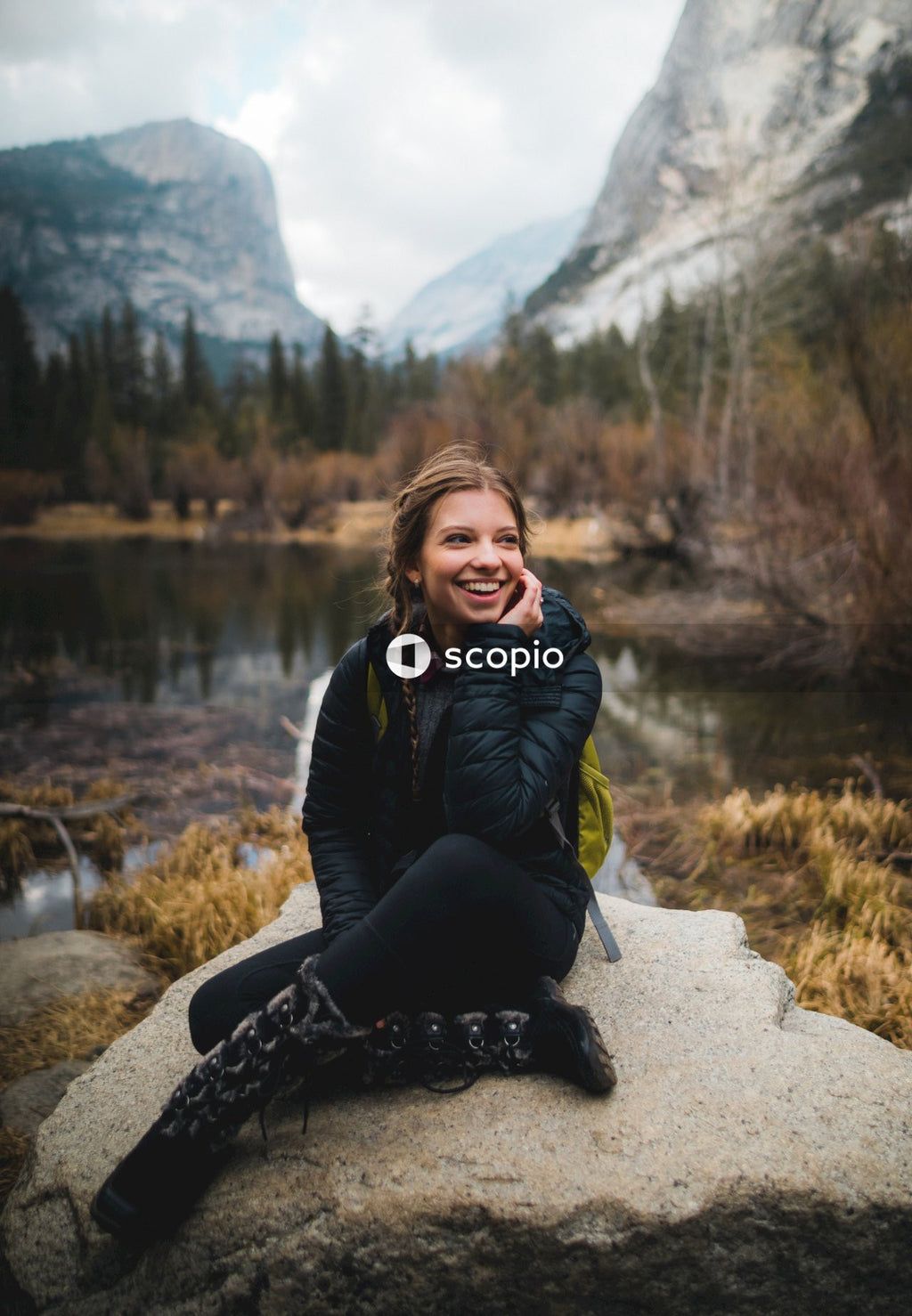 Smiling woman in dark bubble jacket sitting on rock formation near lake