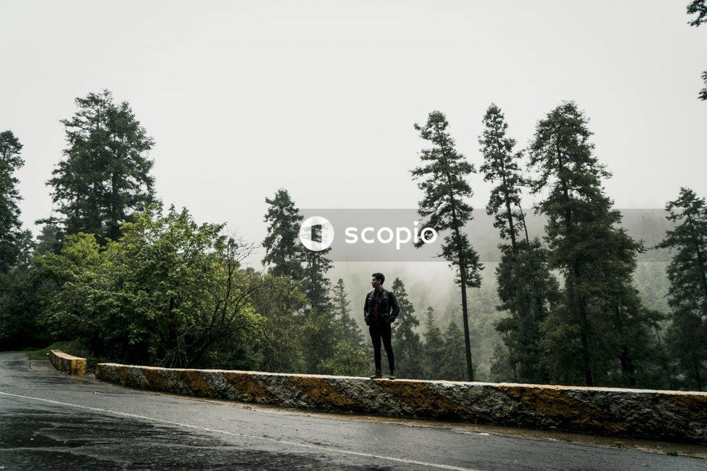 Young man in leather jacket standing on balustrade near pine tree wood