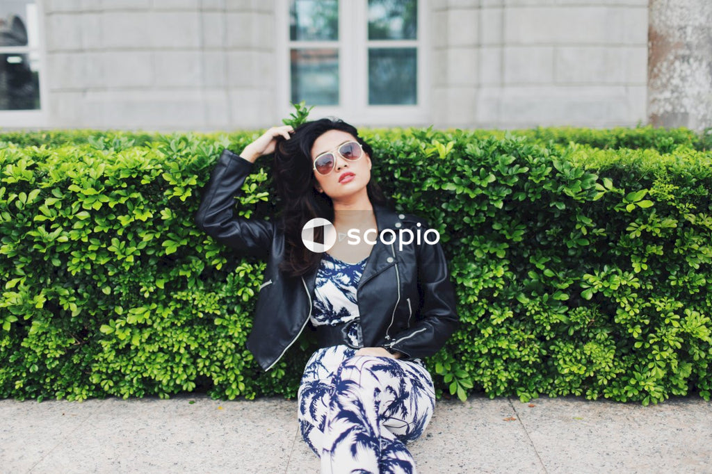 Woman wearing jumpsuit, leather jacket and sunglasses posing on concrete bench in front of bushes