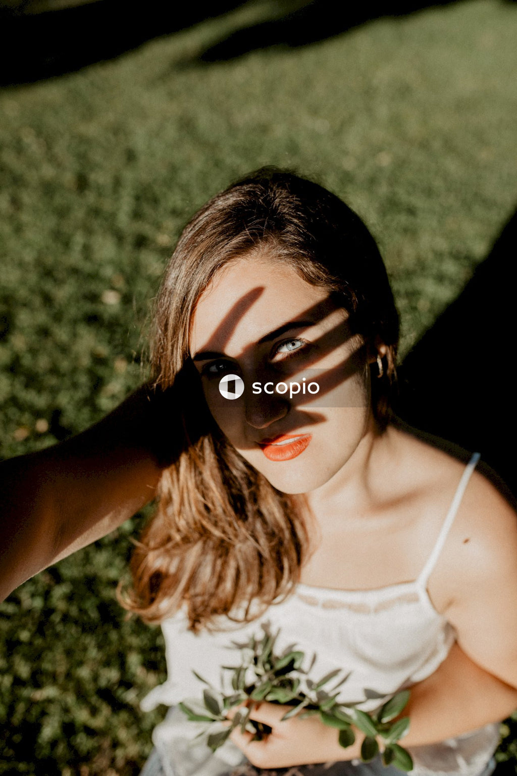Woman in light top with red lipstick standing on green grass blocking sunlight with her hand
