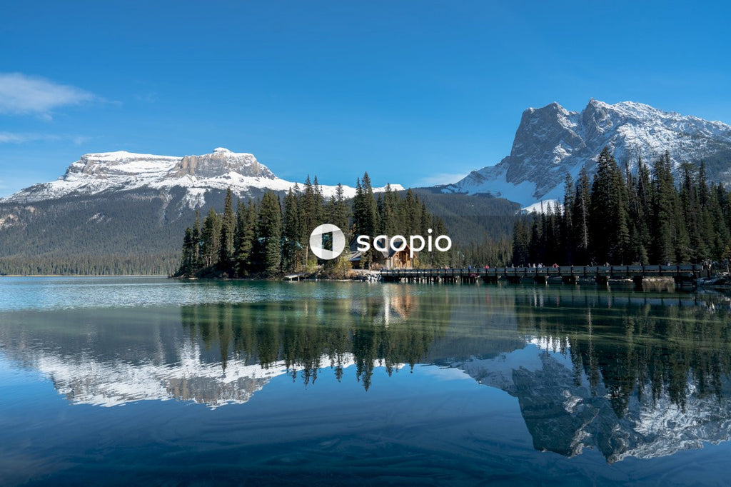 Brown wooden house on lake near green trees and snow covered mountain with reflection on water