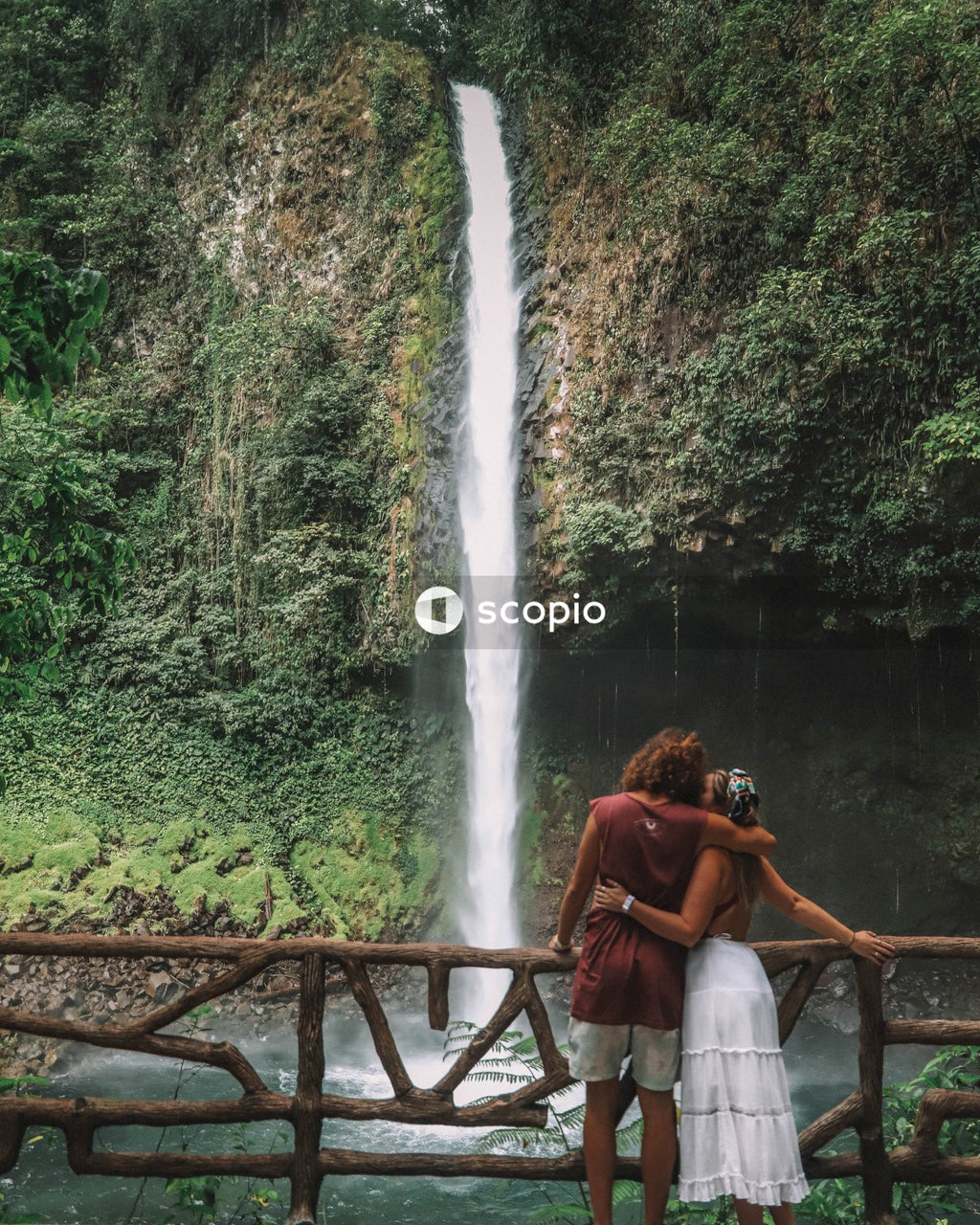 Man and woman standing on brown wooden bridge near waterfalls