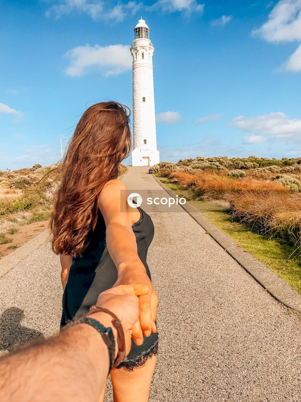 Woman holding a hand of another person and walking toward white lighthouse