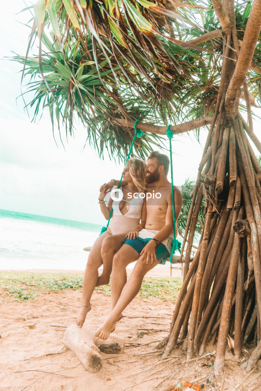 Man and woman in swimsuits sitting on swing chair hanging from tree on beach