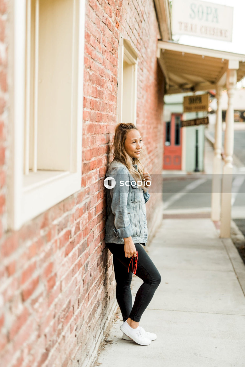 Woman in blue denim jacket and dark pants standing beside brick wall