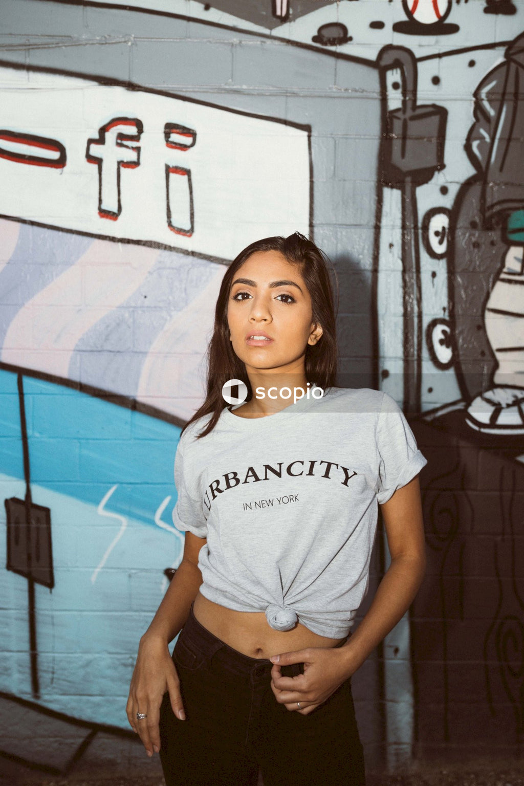 Woman, dressed in a white crop top t-shirt, strikes a pose against a graffiti-covered wall