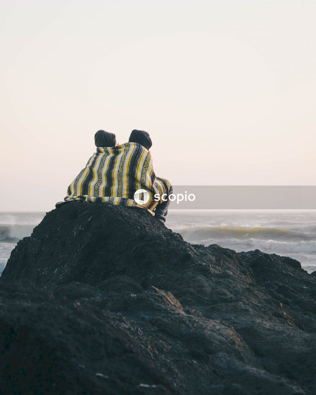 Back view of two people covered with one blanket sitting on a rocky seashore at sunset