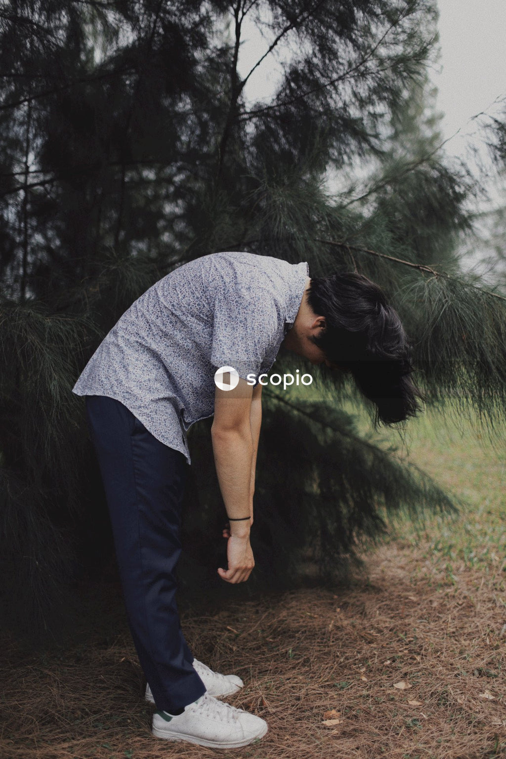 Man standing beside the tree while looking down on his shoes