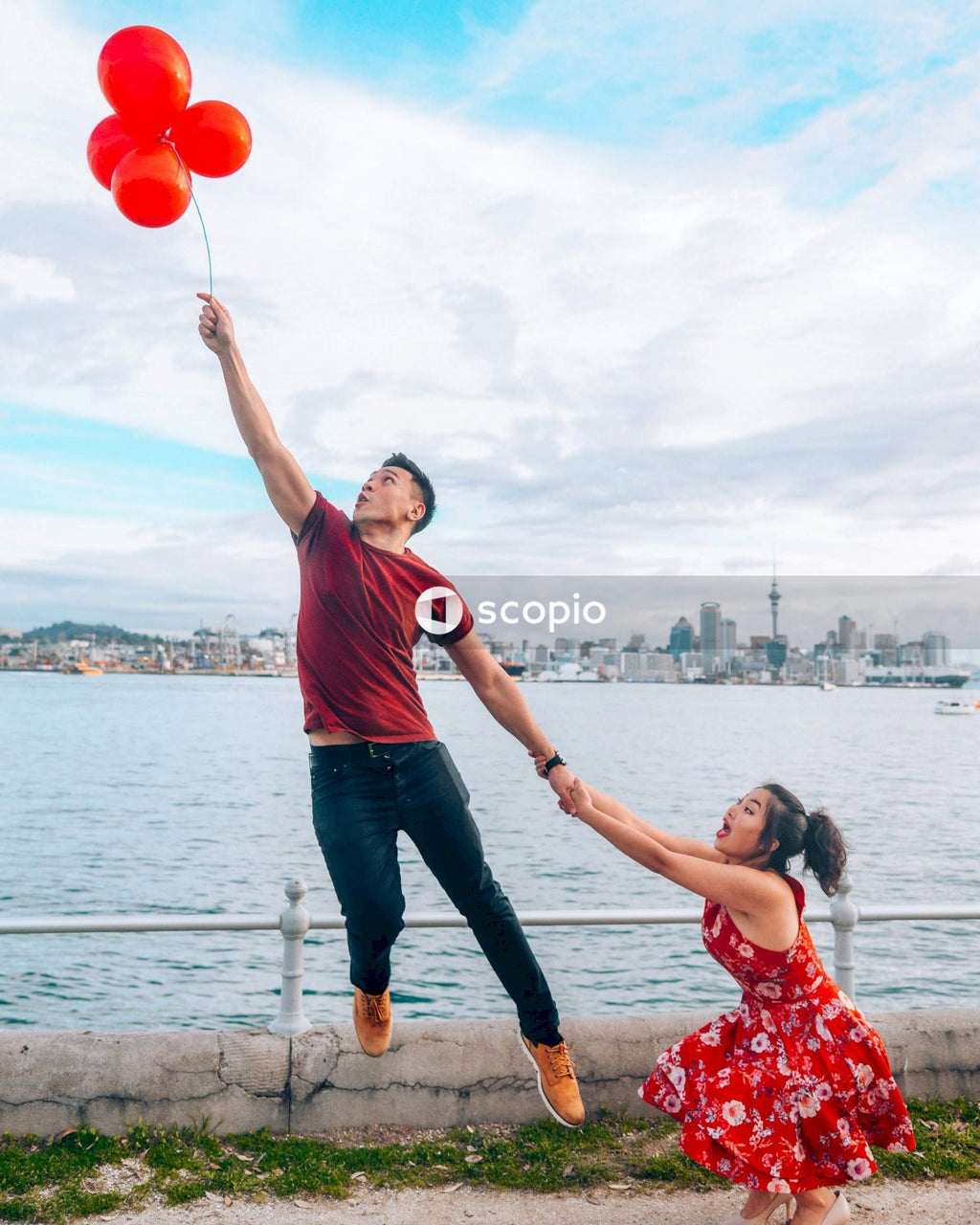 Man in red t-shirt and blue denim jeans holding balloons while a lady is pulling his arm