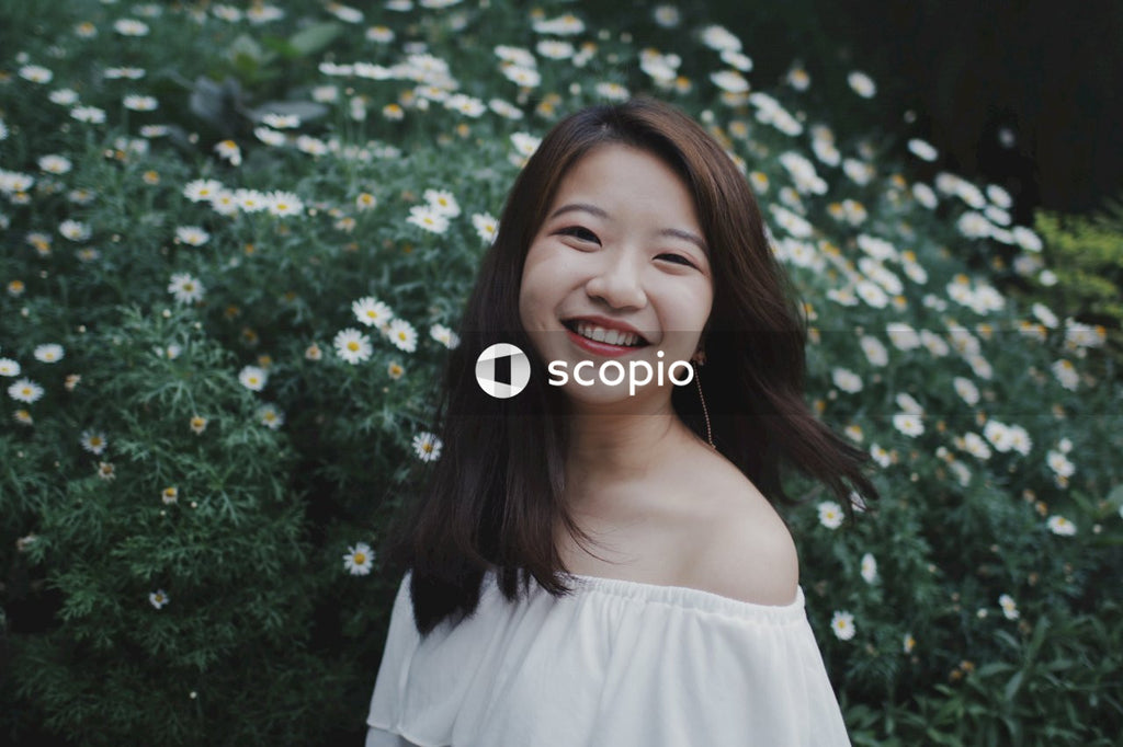 Asian young lady wearing light off-shoulder top with greenery and white flowers behind her smiling