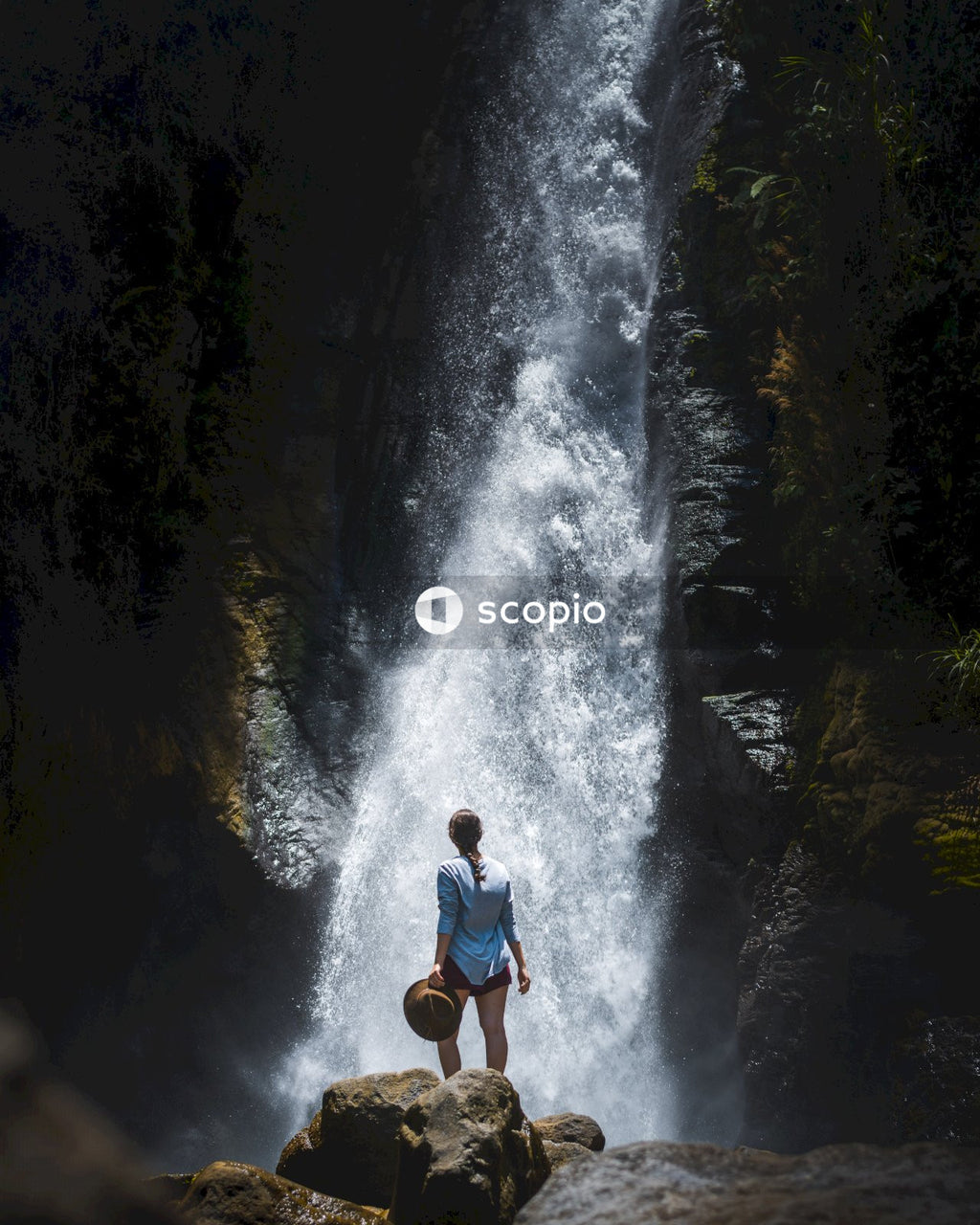 Woman holding a hat and standing on rock near waterfall