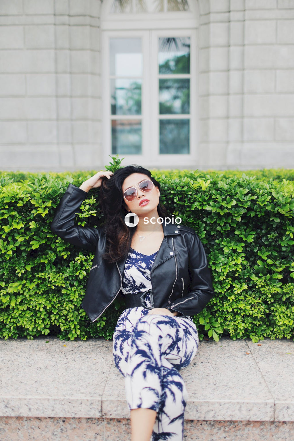 Woman wearing jumpsuit, leather jacket and sunglasses posing on concrete bench in front of bushes