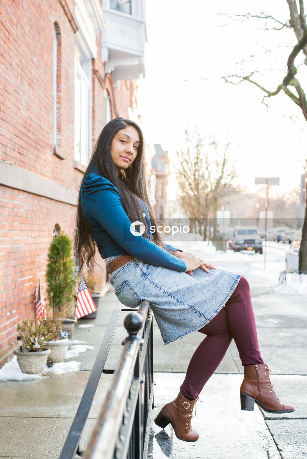Woman in blue long sleeve shirt and denim skirt sitting on gray metal railings on sidewalk