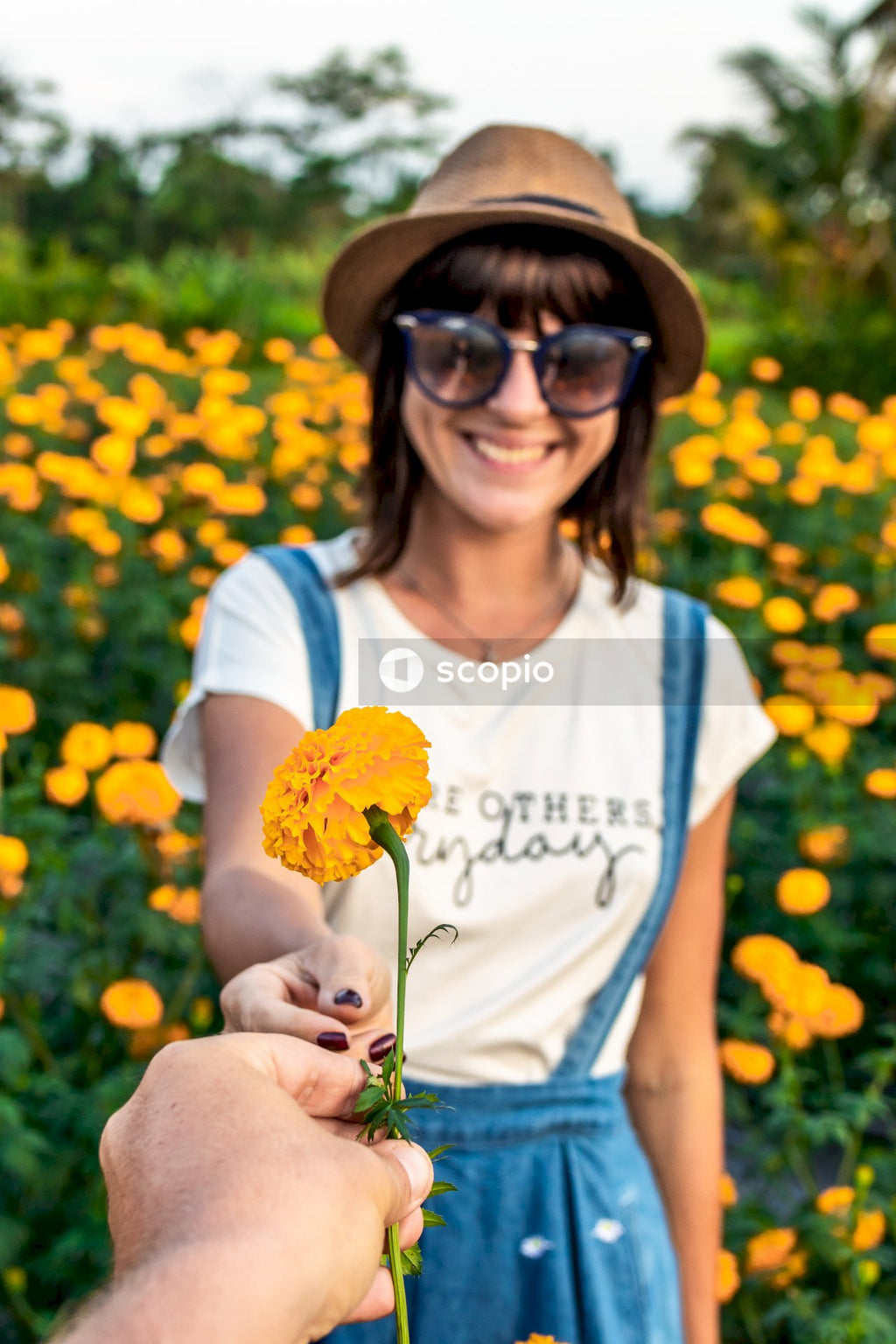 Person giving yellow marigold flower to a woman with brown hat and sunglasses
