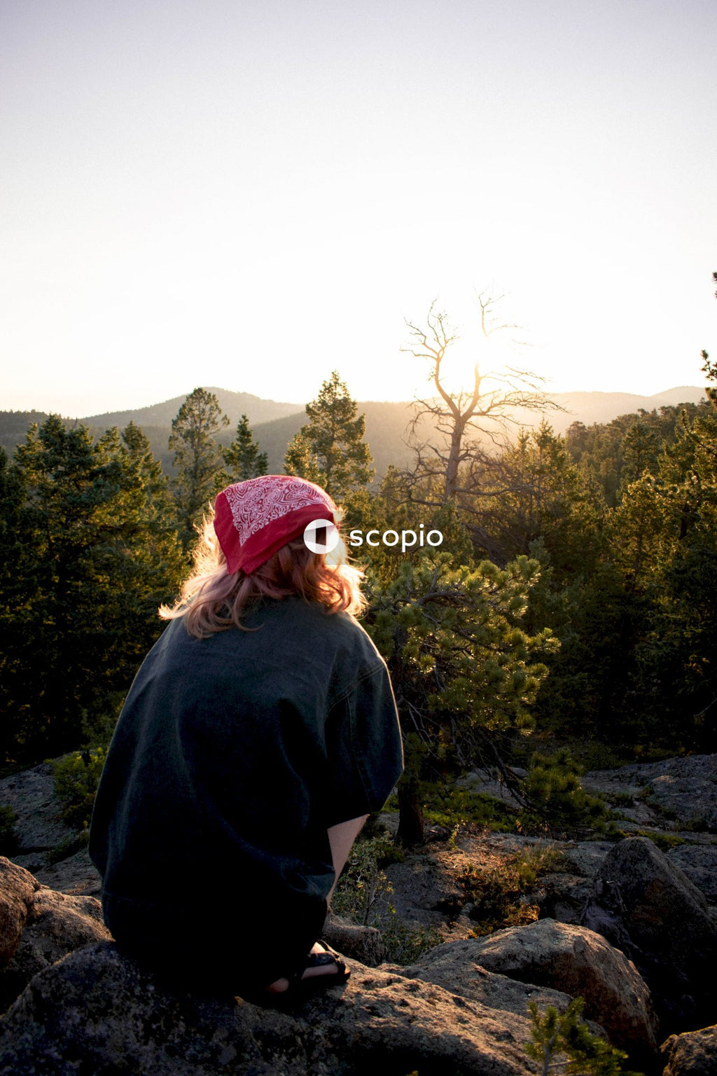 Back view of blonde woman with red bandana crouching on a rock overlooking forest