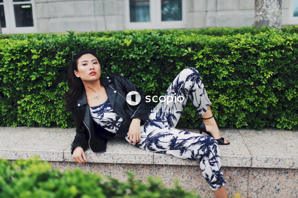 Woman wearing jumpsuit, leather jacket and sunglasses posing on concrete bench in front of bushes