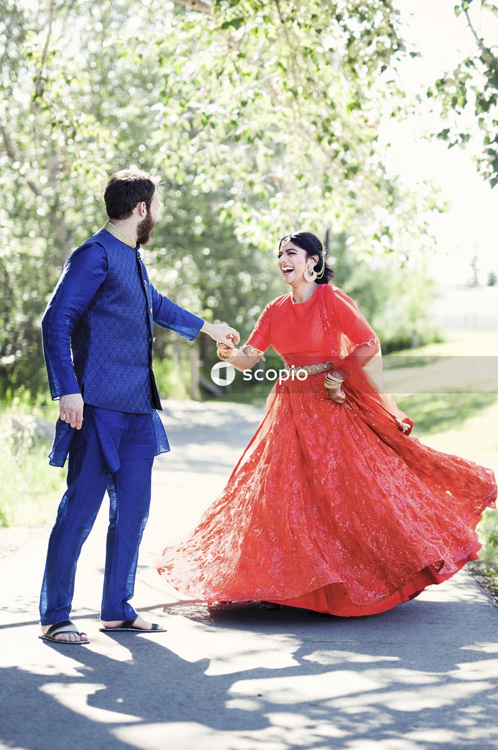 Bride in red sari and groom in blue suit posing near trees