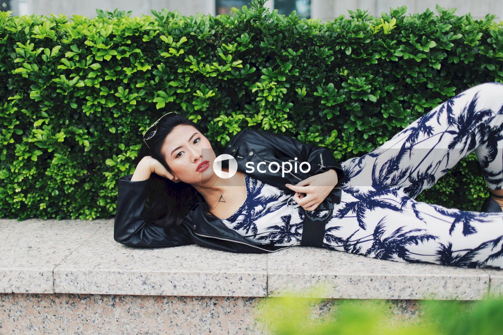 Woman wearing jumpsuit, leather jacket and sunglasses posing on concrete bench in front of bushes