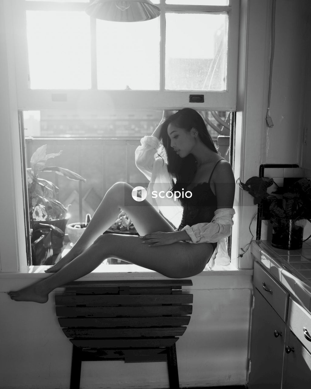 Grayscale photo of woman in lingerie and shirt sitting at a window in the kitchen