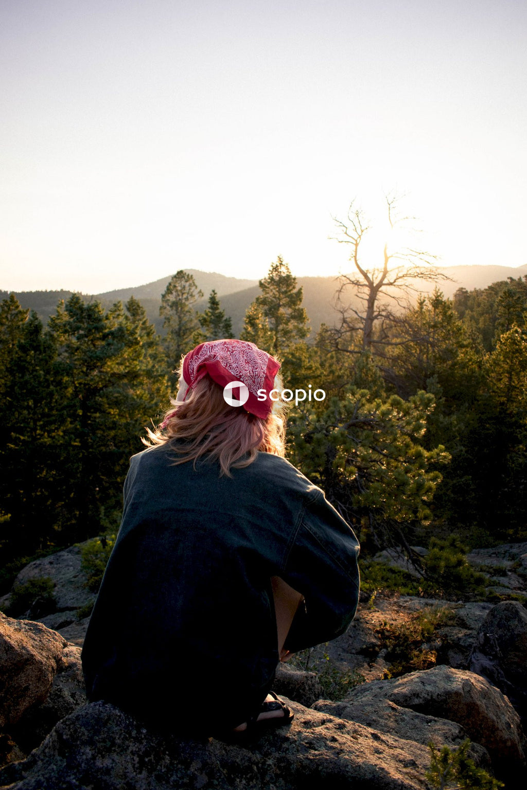 Back view of blonde woman with red bandana crouching on a rock overlooking forest
