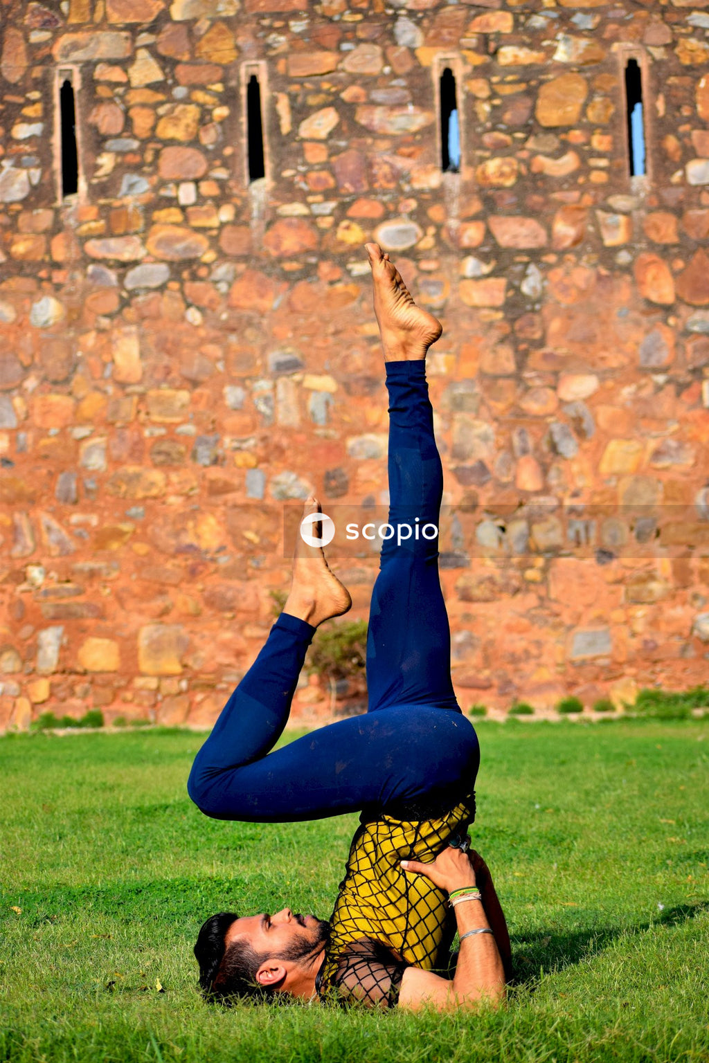 Man doing a shoulder stand on green grass beside brick wall outdoor during daytime