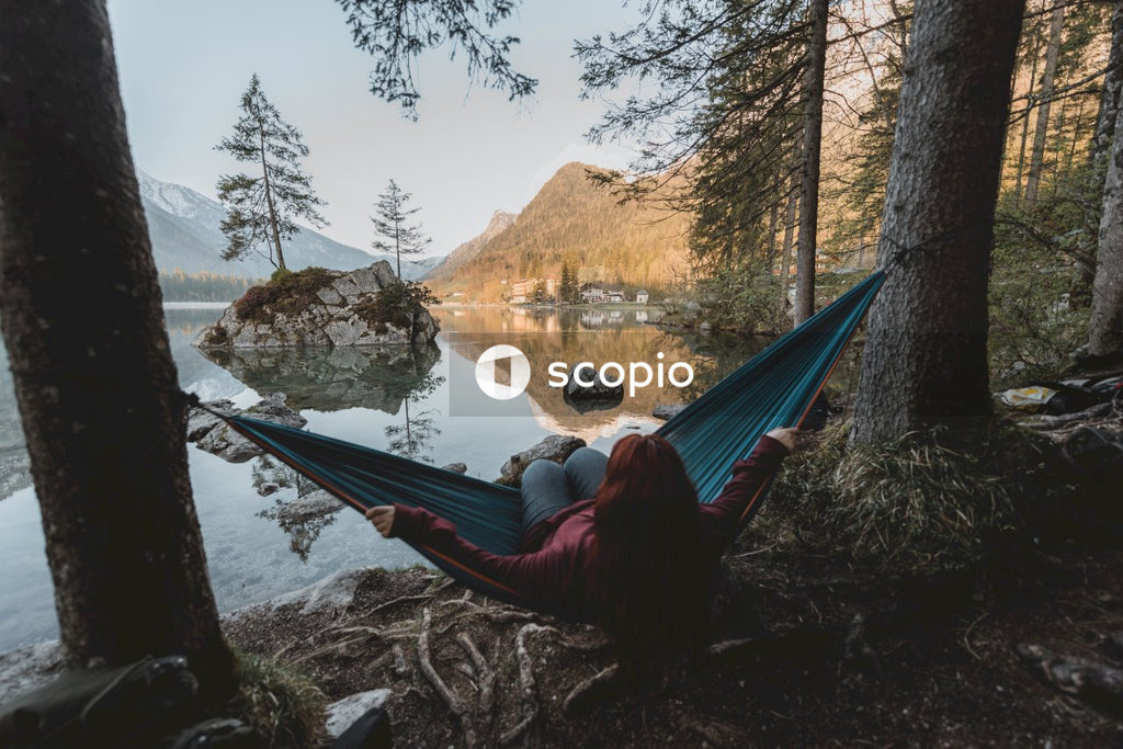 Woman in red jacket sitting on blue hammock near lake during daytime
