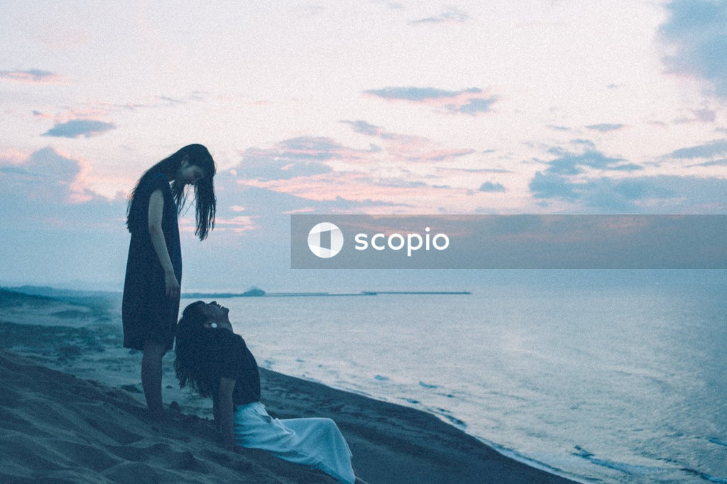 Side view of woman standing behind woman sitting on seashore at sunset