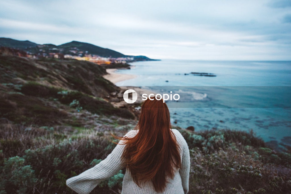 Red haired woman in gray sweater standing on hilltop facing body of water