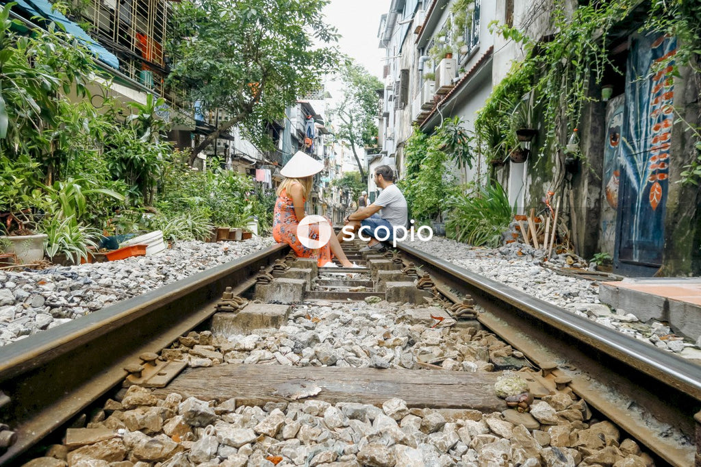 Woman in floral dress wearing conical hat and a man sitting on train rail between houses