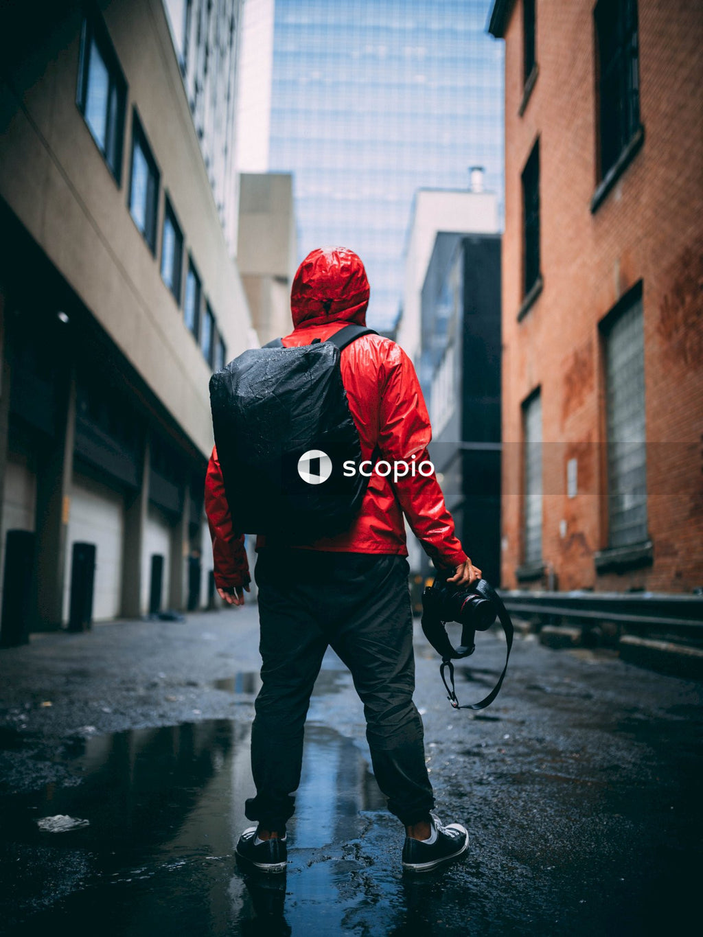 Back view of man in red jacket with backpack holding a camera and standing between buildings