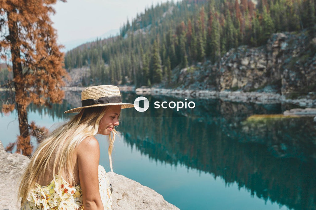 Woman in white floral dress wearing brown straw hat standing on rock near lake