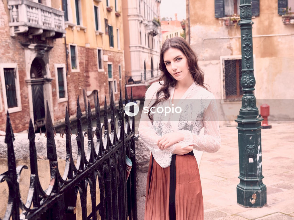 Woman in white lace long sleeve shirt and orange skirt standing beside black metal fence