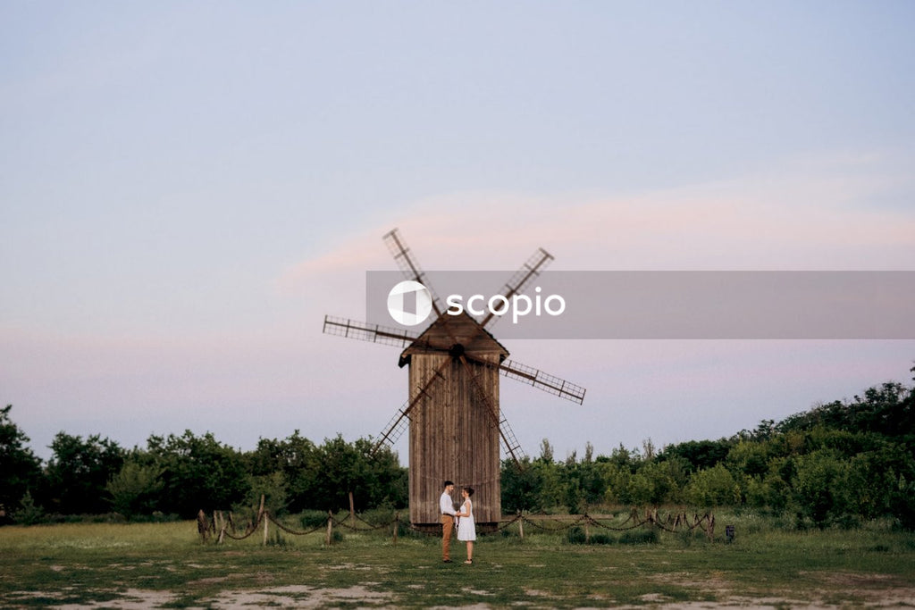 Man and woman holding hands and standing beside wooden windmill on green grass field