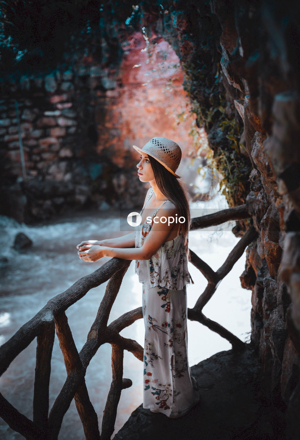 Woman in life floral dress and hat standing beside wooden balustrade on a cliff beside river