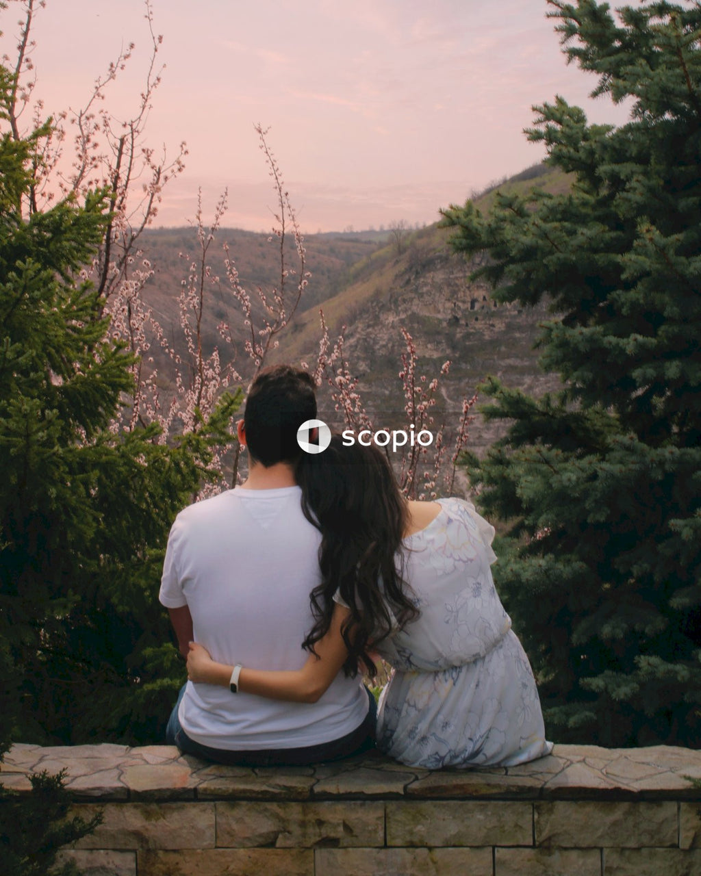 Couple sitting on concrete fence looking at green trees