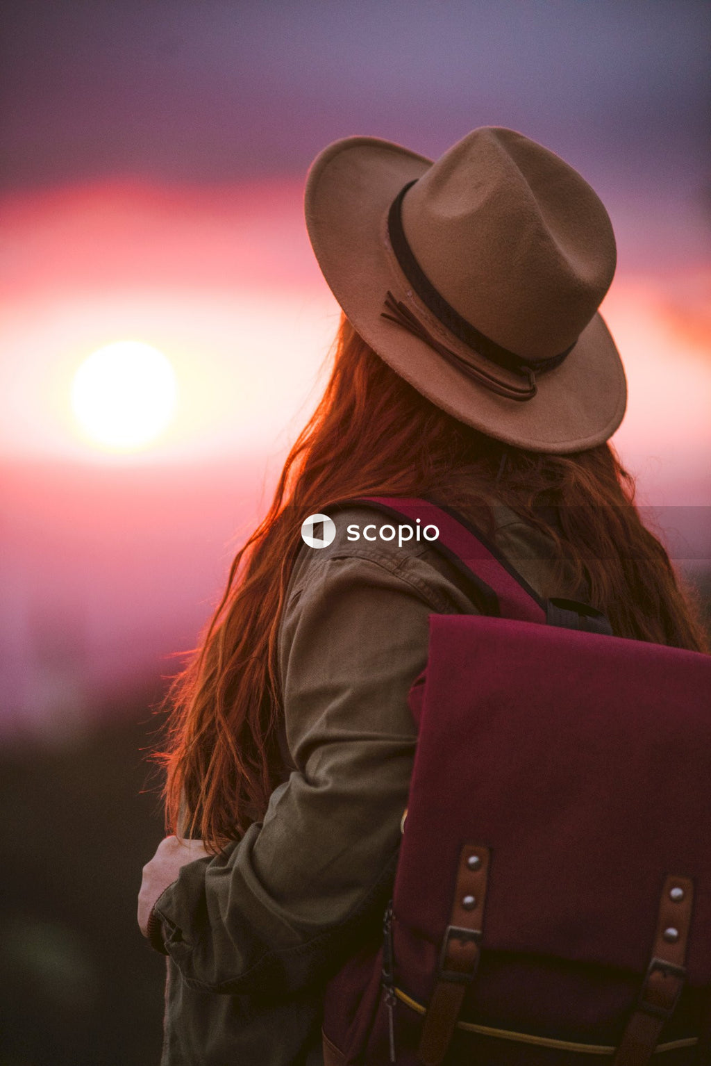 Back view of woman with red hair wearing a hat standing against sunset sky