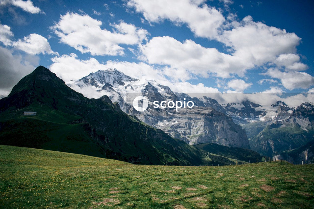 Green grass field near snow covered mountain under white clouds and blue sky
