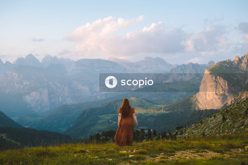 Back view of woman in brown dress standing on grass field against mountainous landscape