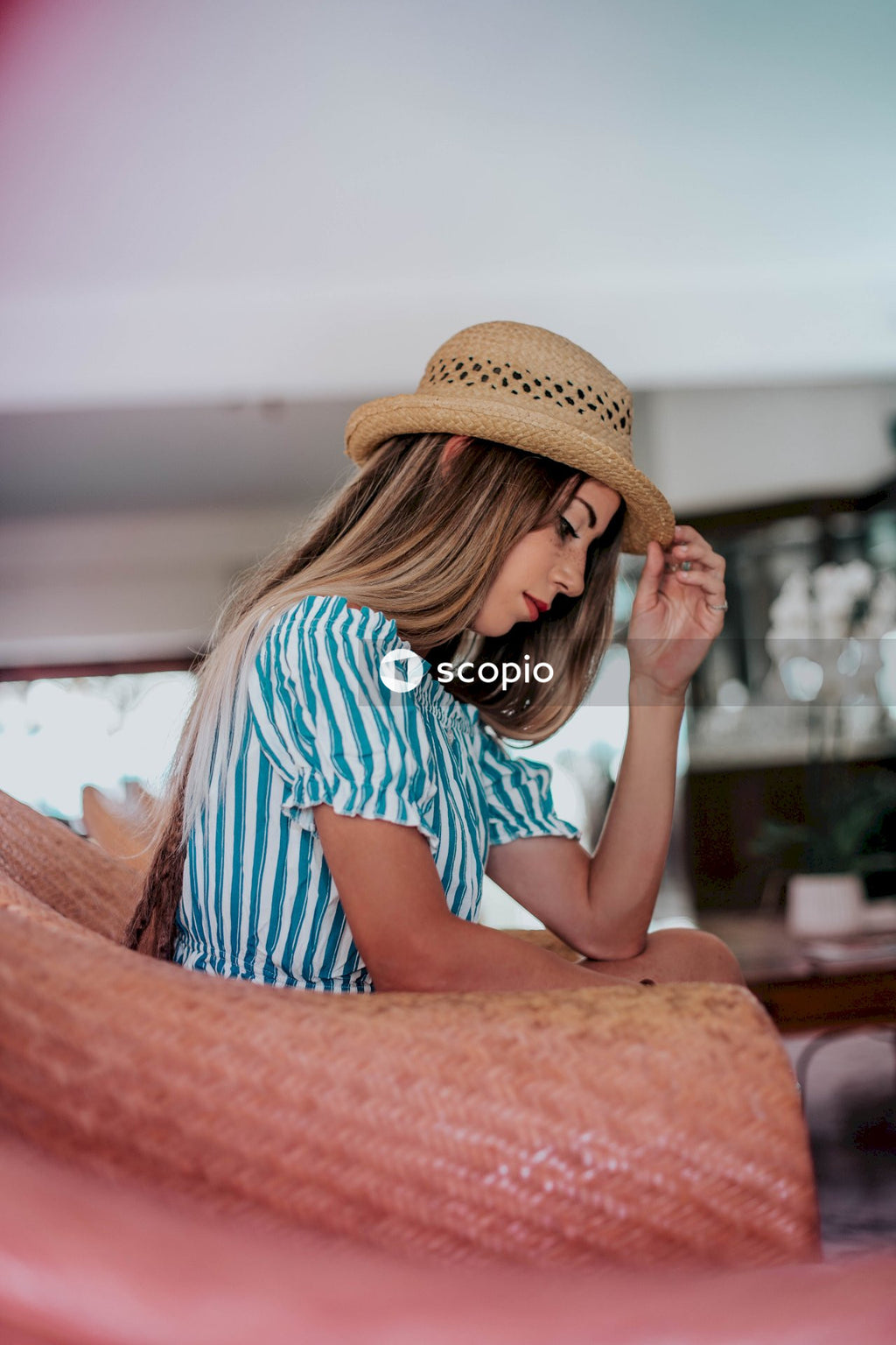Woman in blue and white stripe shirt wearing brown hat sitting pink armchair indoor
