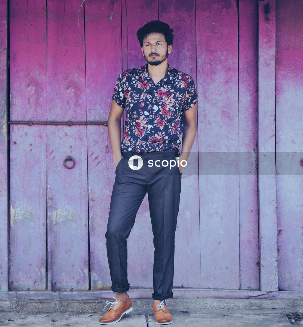 Man in black and white floral shirt and black pants standing beside red wooden door