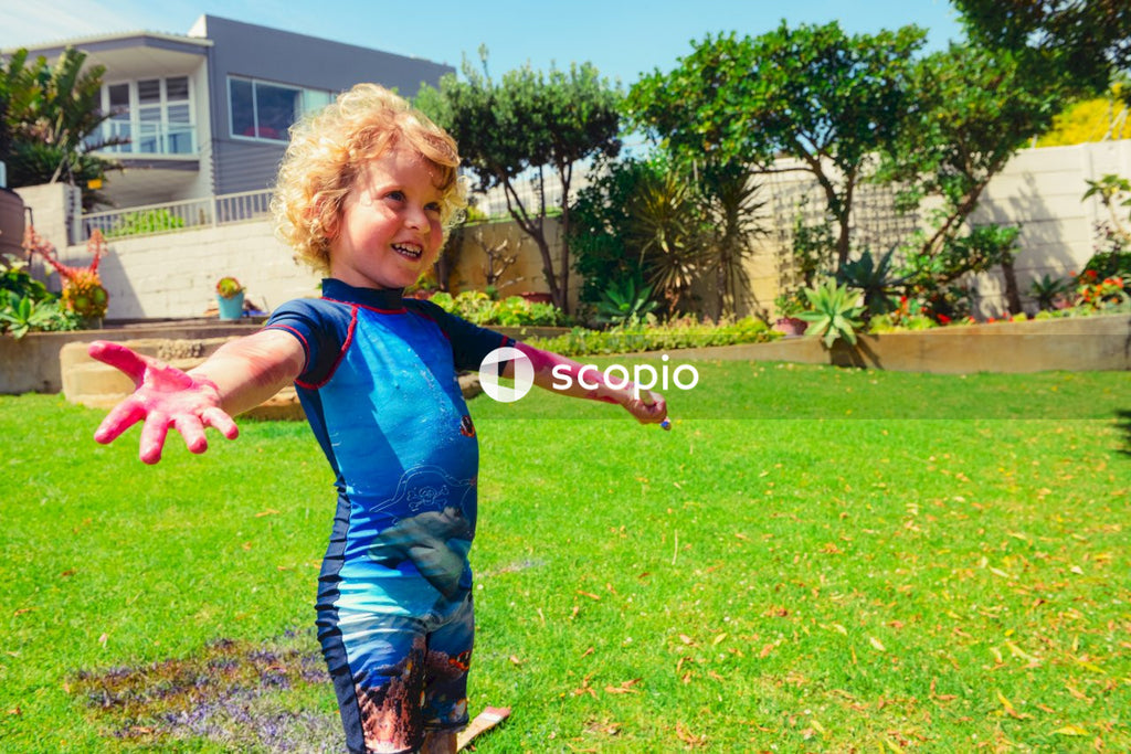 Blond boy in a summer costumes standing on green grass lawn with opened pink painted hands