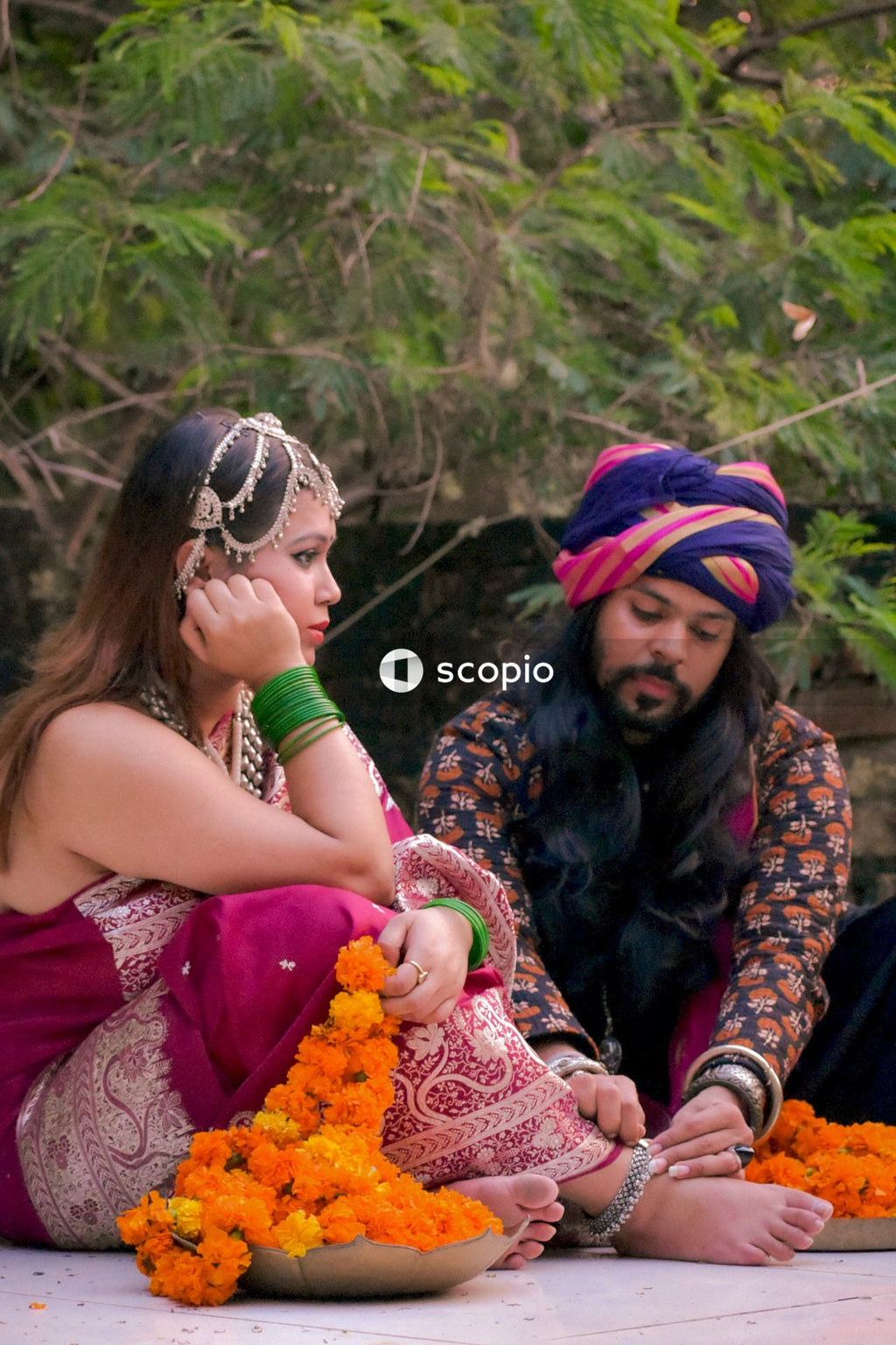 Indian man with long hair wearing turban sitting beside woman in colorful dress on floor outdoor