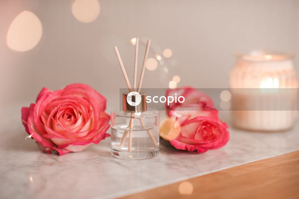 Liquid scented home fragrance in glass bottle and fresh rose flowers over burning candle and glow lights on marble table in room. Cozy atmosphere.