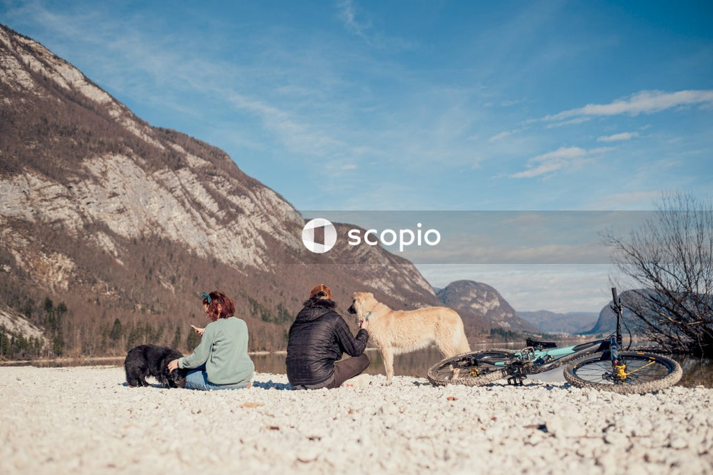 Man and woman sitting on sand near brown rock mountain with two dogs and a bike
