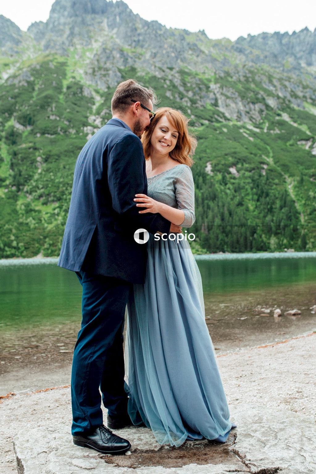 Man in black long sleeve shirt hugging woman in gray dress near body of water