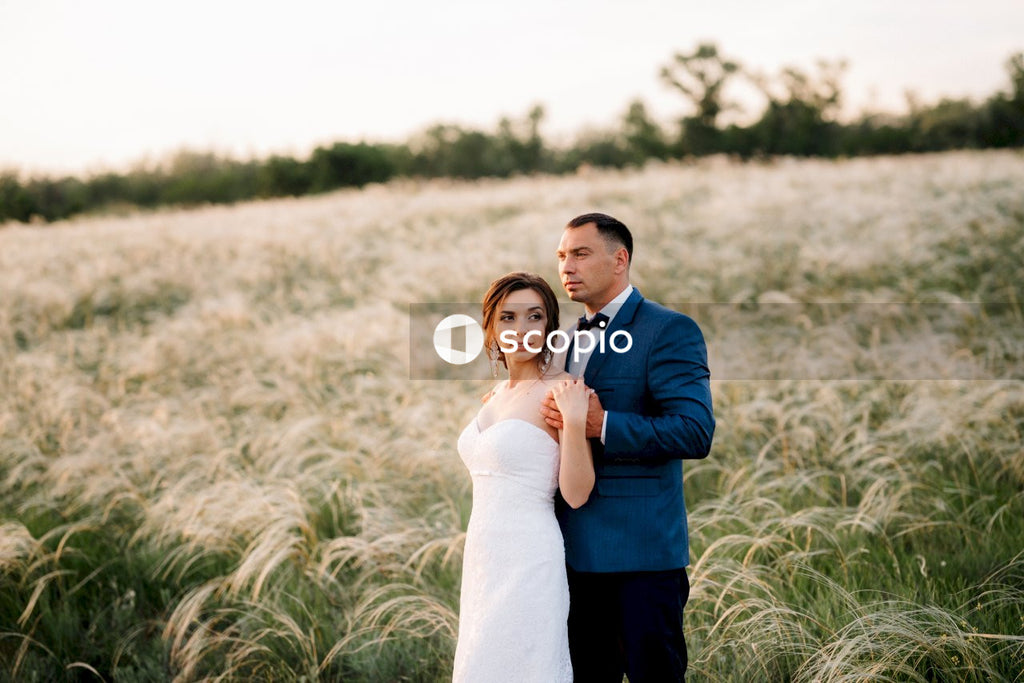 Man in black suit jacket hugging woman in white dress on green grass field