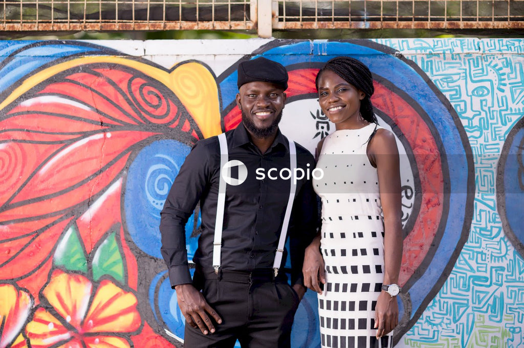 Man in dark shirt with strap and hat and a woman in light dress smiling against a graffiti wall