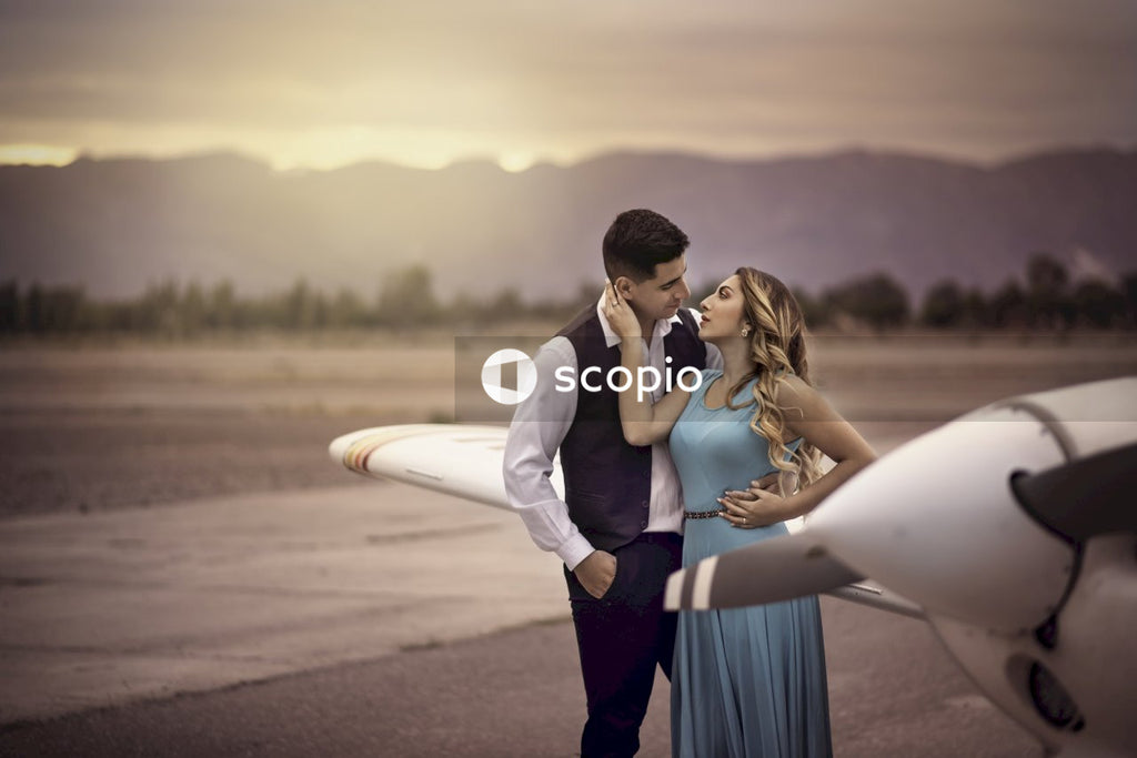 Man in dark vest kissing woman in blue dress beside an airplane