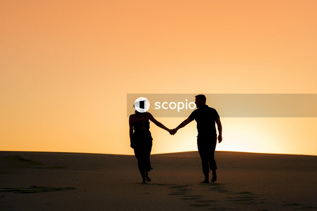 Silhouette of man and woman holding hands standing on sand during sunset