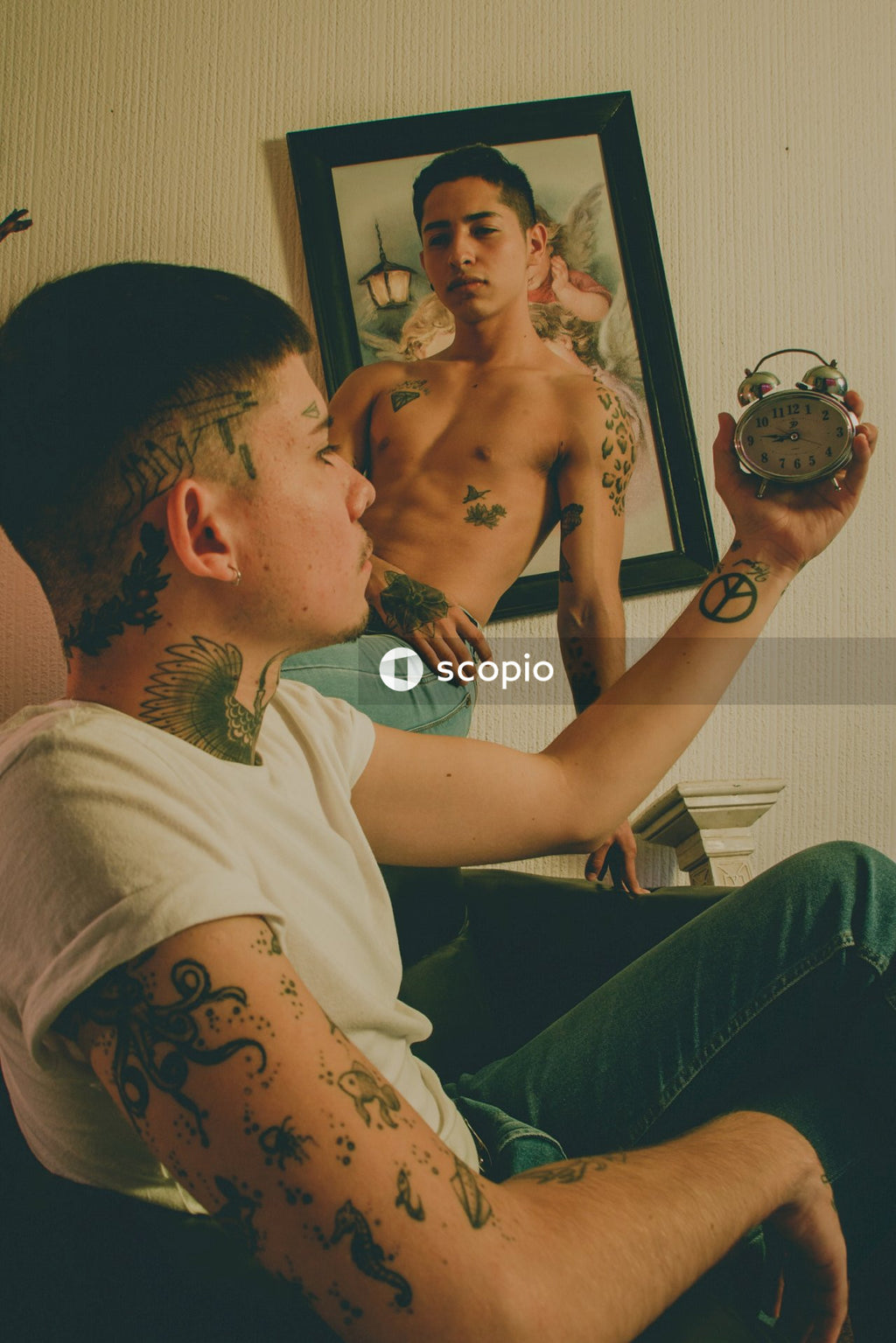 Tattooed man holding an alarm clock while another man sitting and watching